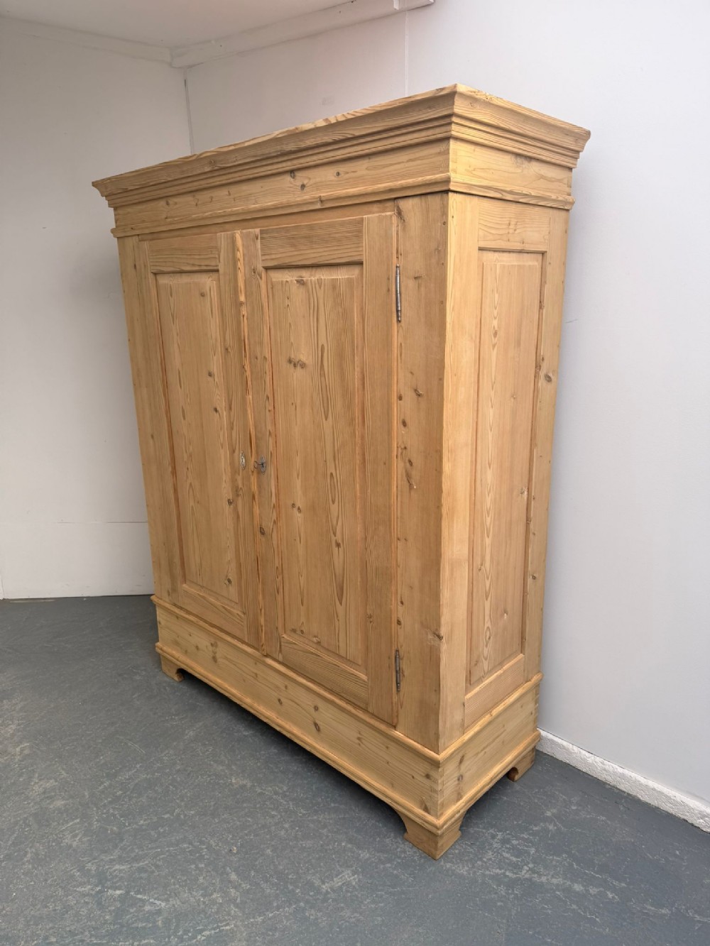 A freestanding natural pine two-door wardrobe featuring panelled doors, a stepped pediment cornice, and a plinth base.