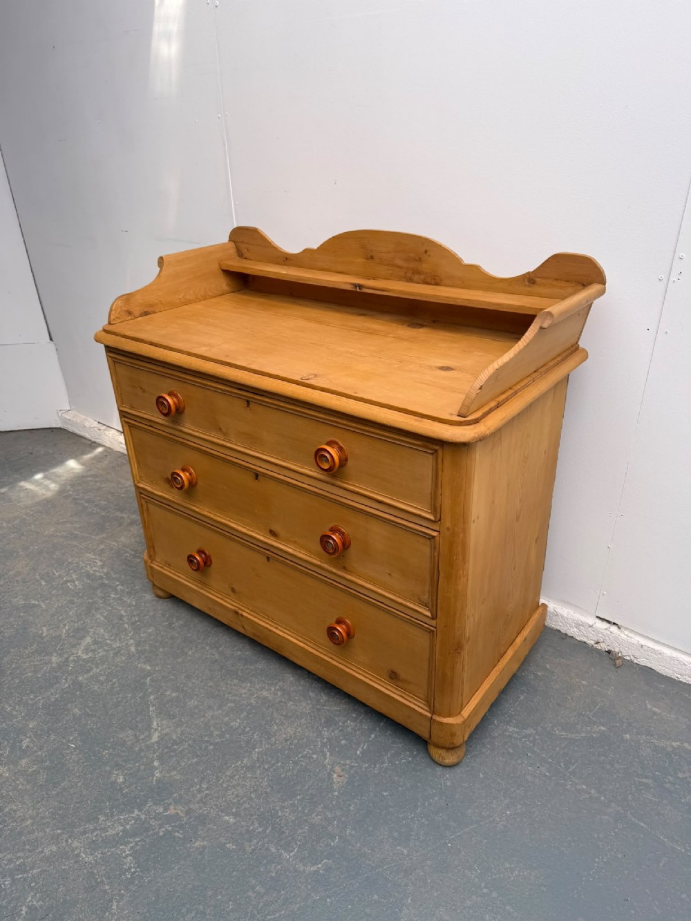 An early Victorian waxed pine chest of drawers featuring three wide drawers with turned wooden knobs and a shaped gallery upstand.