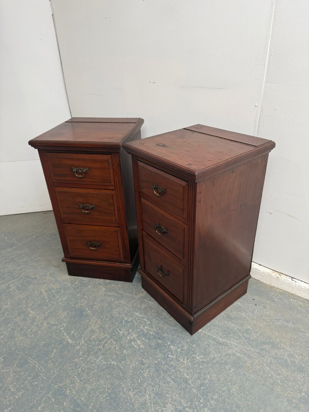 A pair of narrow mahogany bedside chests featuring three drawers each with brass handles on a plinth base.