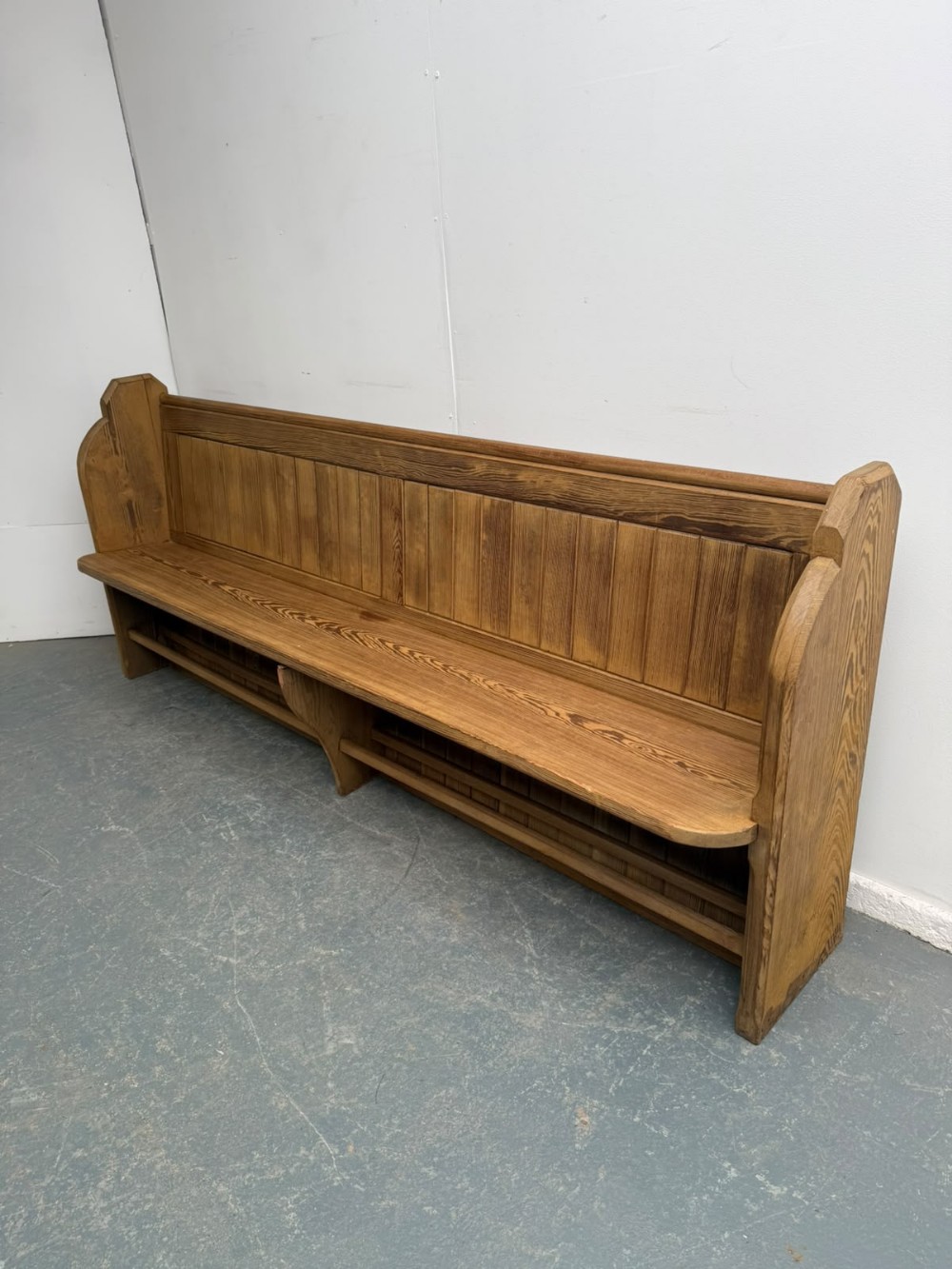 A long Victorian pine church pew featuring a vertical panelled backrest, shaped end supports, and an under-seat rack.