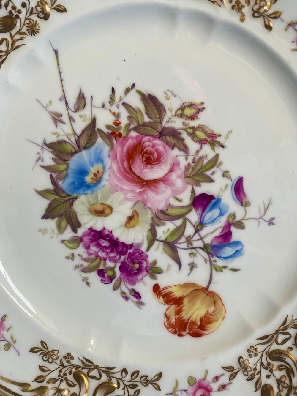 A circular white soft-paste porcelain plate featuring a hand-painted central floral bouquet and a scalloped rim with ornate gilded rococo scrolling motifs. View II