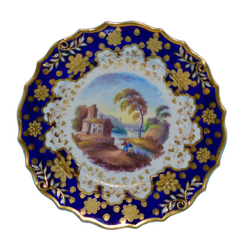 An antique circular porcelain dessert plate featuring a rich cobalt blue rim with gilt floral decoration surrounding a central hand-painted landscape scene.