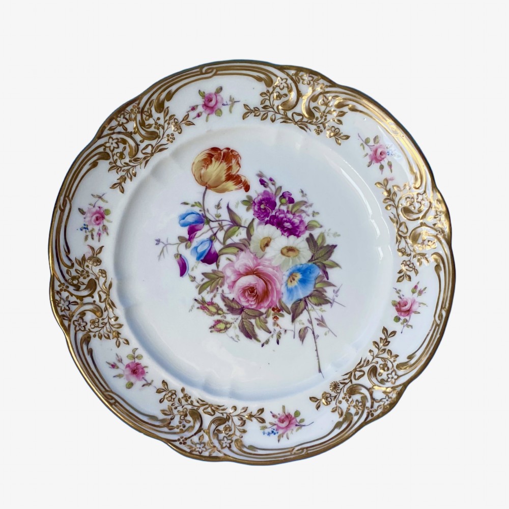 A circular white soft-paste porcelain plate featuring a hand-painted central floral bouquet and a scalloped rim with ornate gilded rococo scrolling motifs.