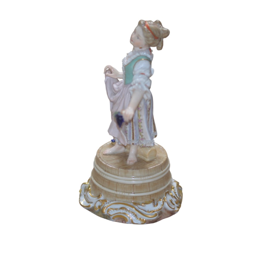 A polychrome Meissen porcelain figurine depicting a female figure holding grapes atop a circular pedestal with gilded scrollwork.