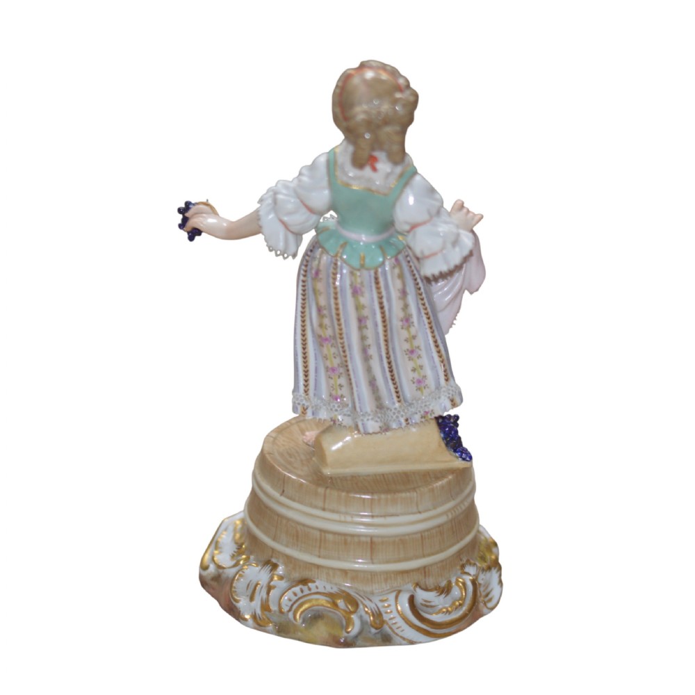 A polychrome Meissen porcelain figurine depicting a female figure holding grapes atop a circular pedestal with gilded scrollwork. View II