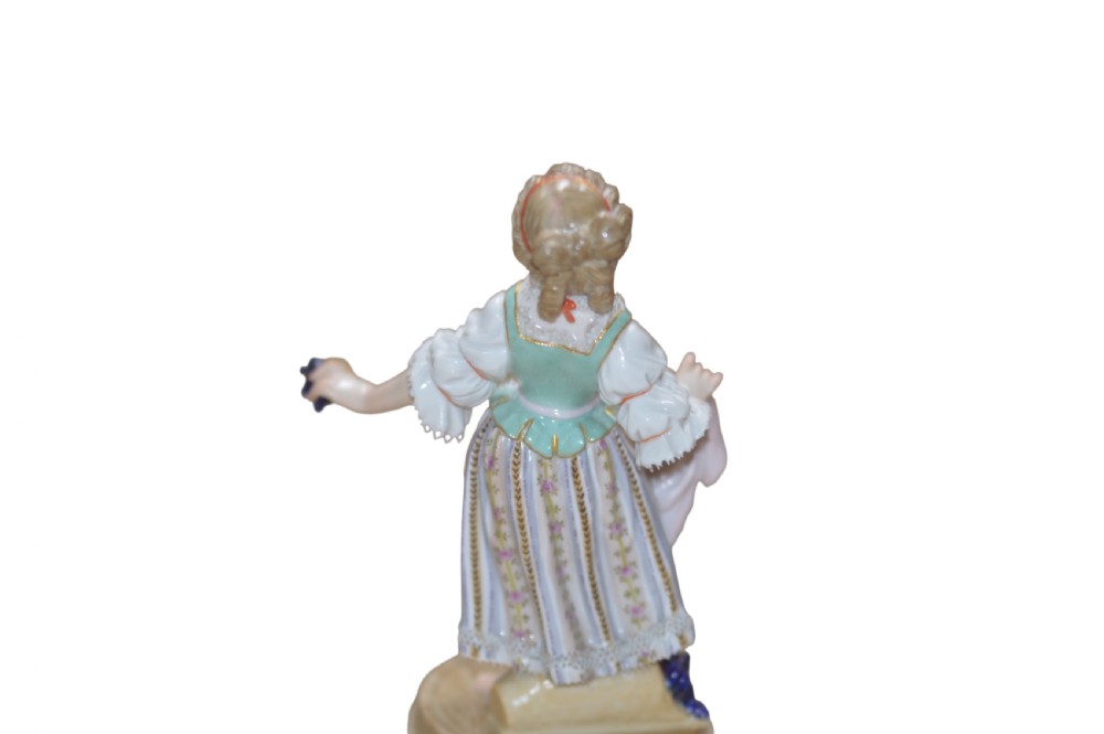 A polychrome Meissen porcelain figurine depicting a female figure holding grapes atop a circular pedestal with gilded scrollwork. View VI