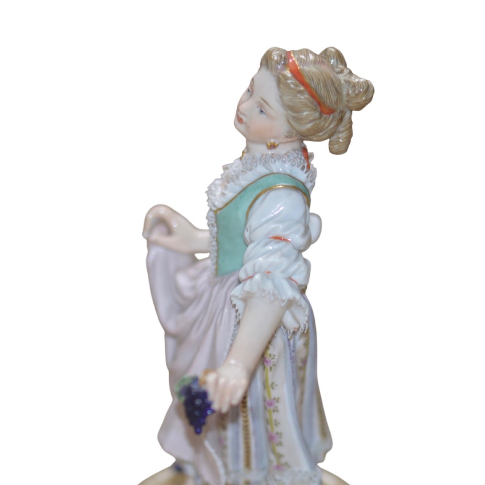 A polychrome Meissen porcelain figurine depicting a female figure holding grapes atop a circular pedestal with gilded scrollwork. View XII
