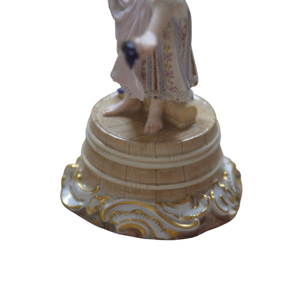A polychrome Meissen porcelain figurine depicting a female figure holding grapes atop a circular pedestal with gilded scrollwork. View XIII