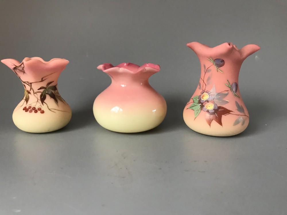 Three small Victorian Burmese glass vases with shaded pink to yellow satin bodies, crimped rims, and hand-painted botanical enamel decorations.
