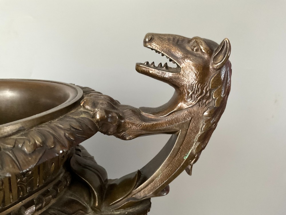 A patinated bronze urn with beast-head handles and a diaper pattern body, mounted on a square red marble plinth. View II