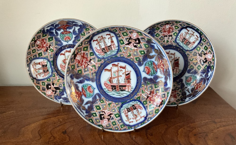A trio of circular Japanese Imari porcelain plates featuring hand-painted European ships, figures, and geometric patterns in cobalt blue, iron red, green, and gold.