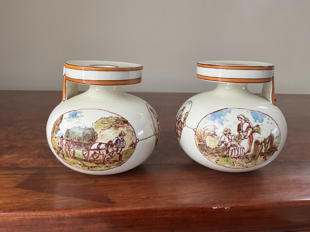A pair of bulbous cream earthenware vases with orange-trimmed tops, decorated with painted oval cartouches depicting rural agricultural harvest scenes.