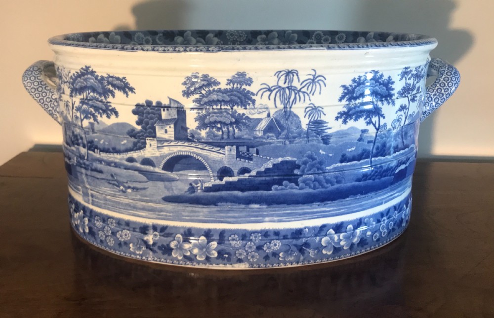 An oval blue and white transferware ceramic footbath featuring twin side handles and a detailed river landscape scene.