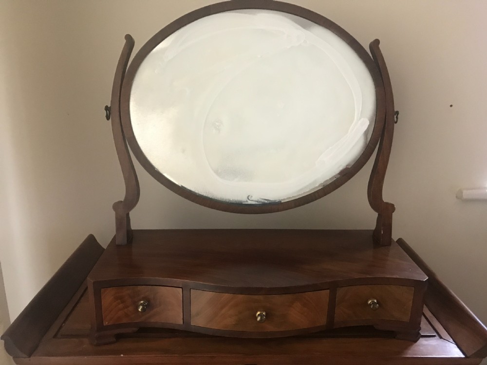 A George III mahogany toilet mirror featuring an oval adjustable glass framed by shaped upright supports above a box base with three small drawers.