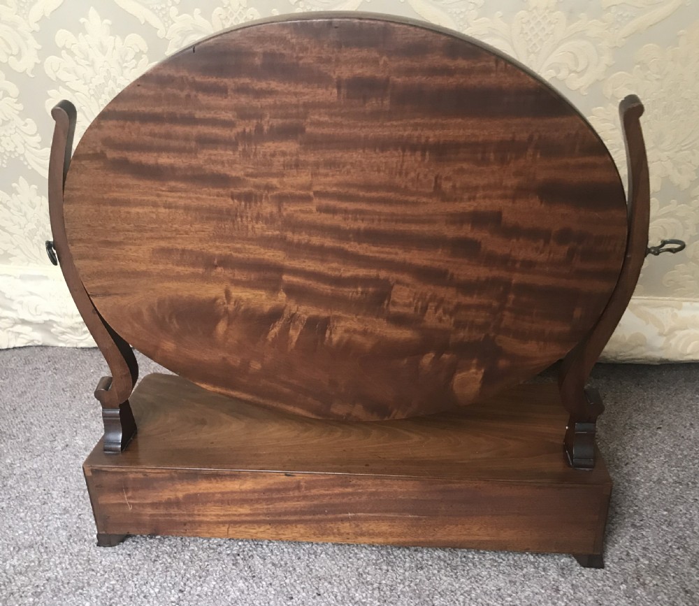 A George III mahogany toilet mirror featuring an oval adjustable glass framed by shaped upright supports above a box base with three small drawers. View II