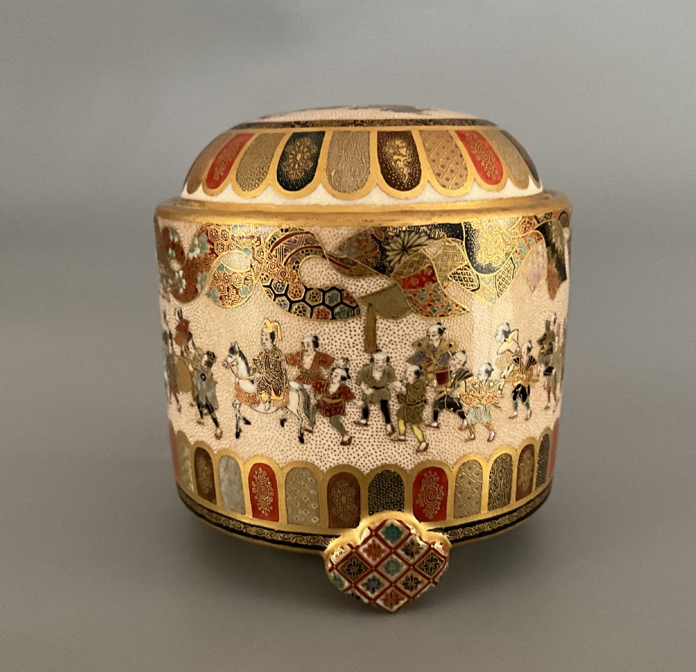 A cylindrical cream earthenware Satsuma incense burner decorated with hand-painted figures in a procession, framed by geometric lappet borders and gold detailing.