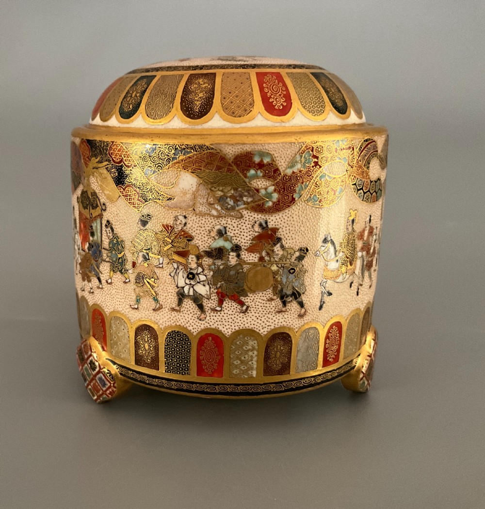 A cylindrical cream earthenware Satsuma incense burner decorated with hand-painted figures in a procession, framed by geometric lappet borders and gold detailing. View II