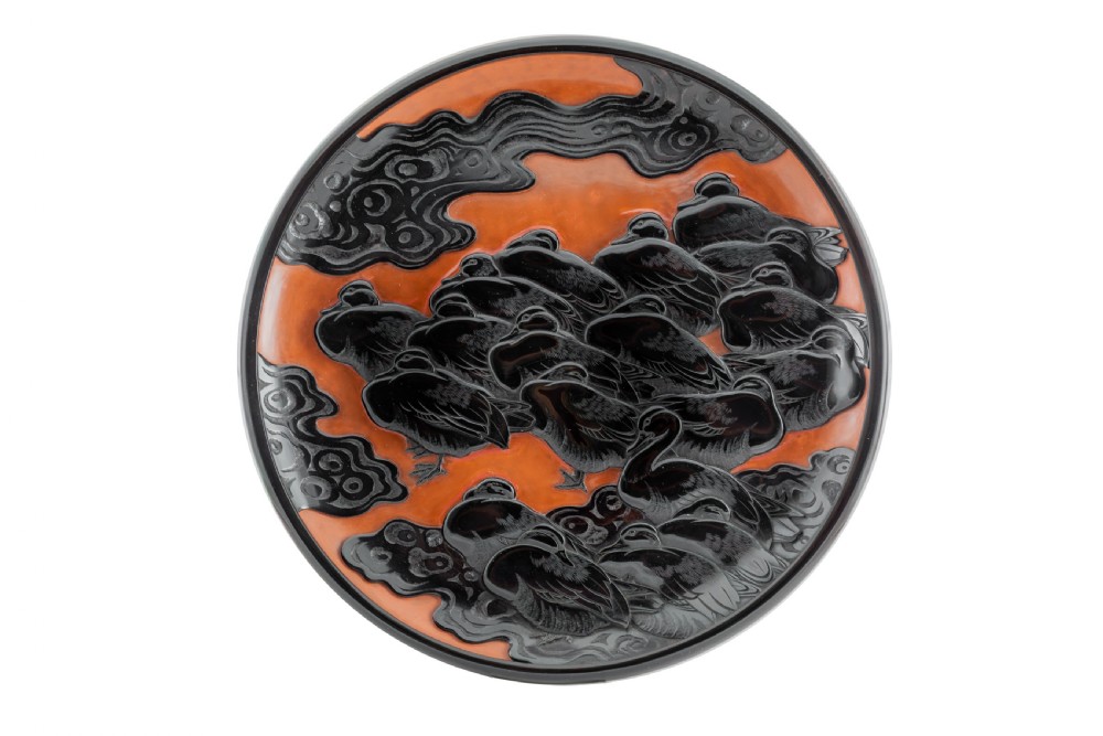 A circular Japanese lacquer plaque featuring carved black water birds and swirling clouds against a textured orange background.