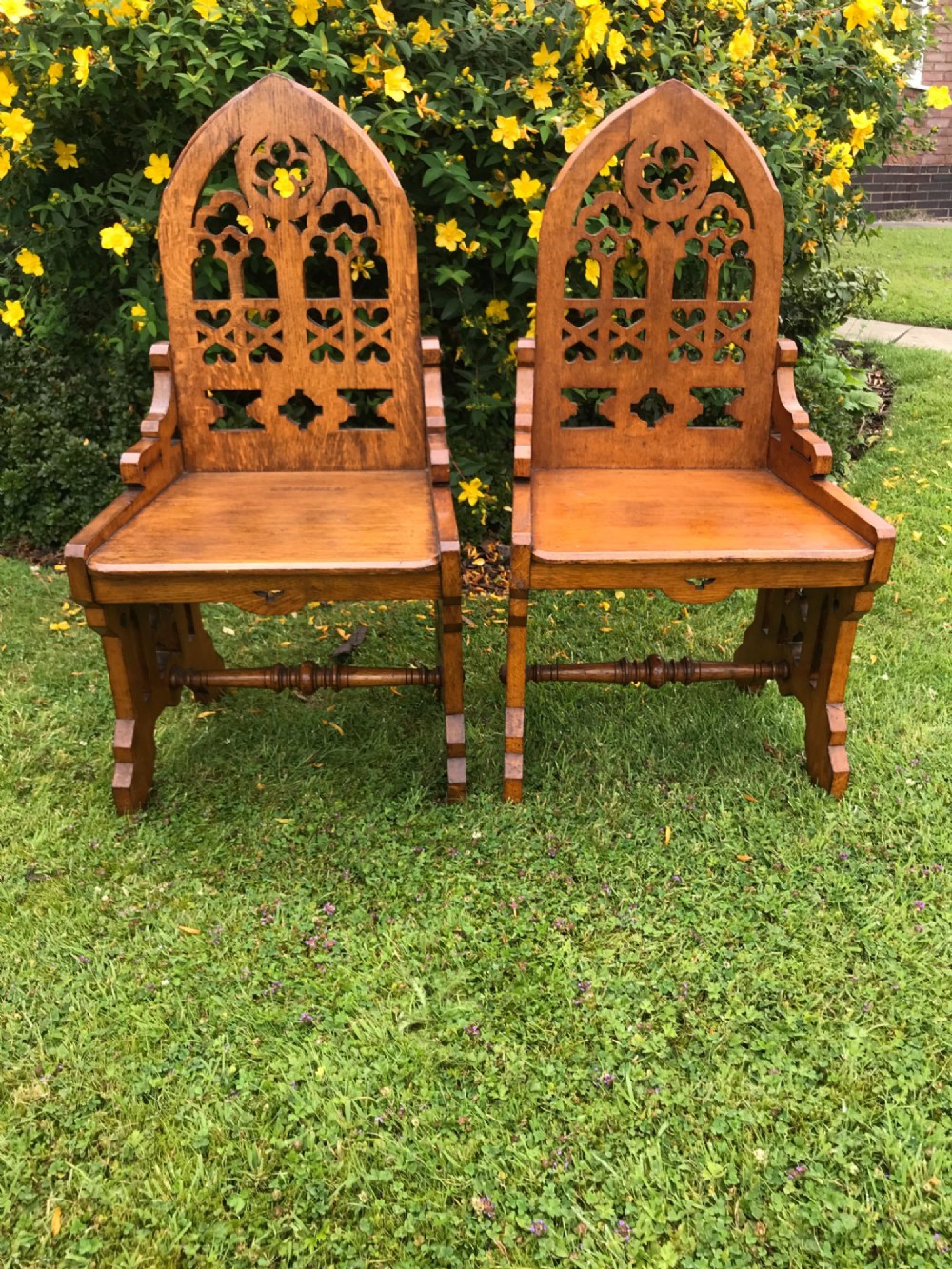 A pair of carved oak Gothic Revival hall chairs featuring pointed arch backs with tracery cut-outs and solid wooden seats.