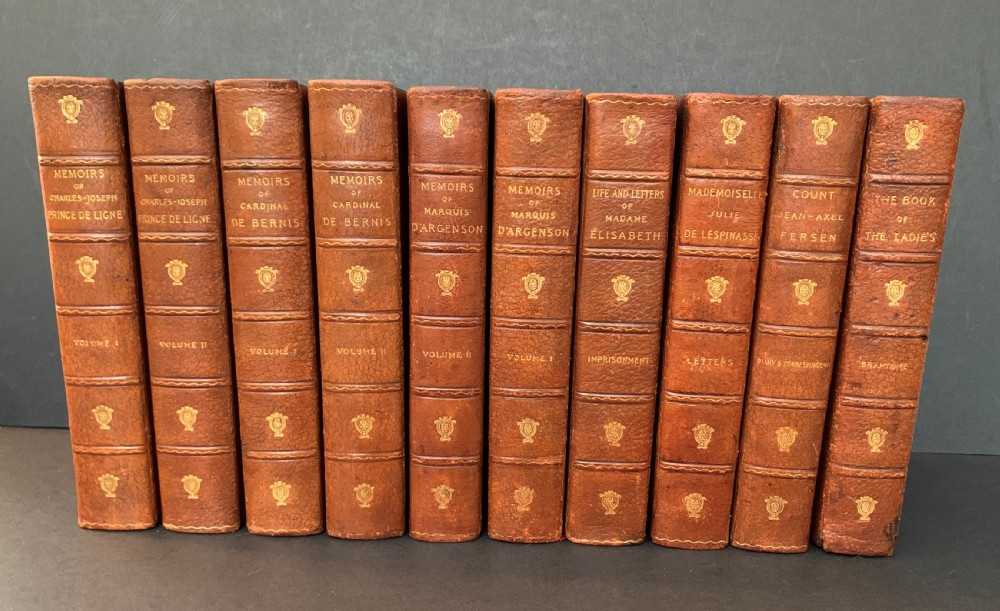 A collection of ten matching antique tan leather-bound books standing upright with gold leaf lettering on their spines.
