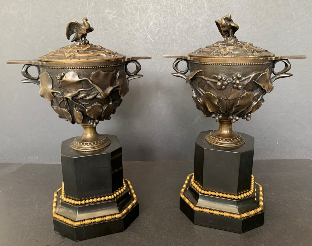 A pair of Victorian cast bronze lidded urns featuring foliage reliefs and bird finials, mounted on octagonal black marble plinths.