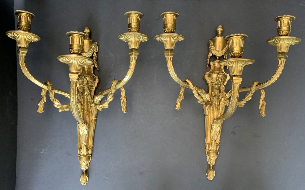 A pair of French gilt bronze wall lights featuring three curving candle branches decorated with leaf swags and topped with urn backplates.