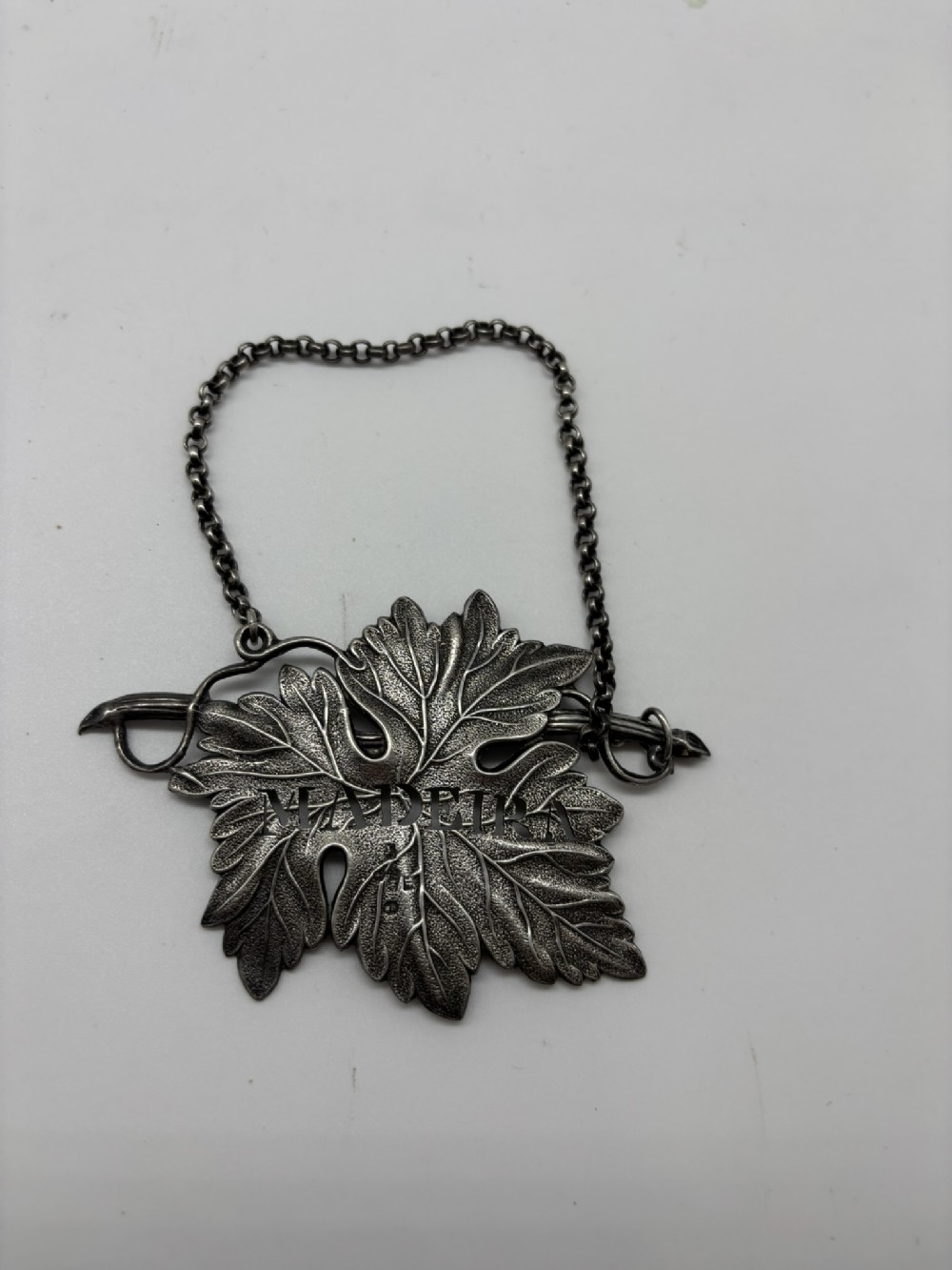 A vine leaf shaped silver decanter label pierced with the word MADEIRA and attached to a silver link chain.