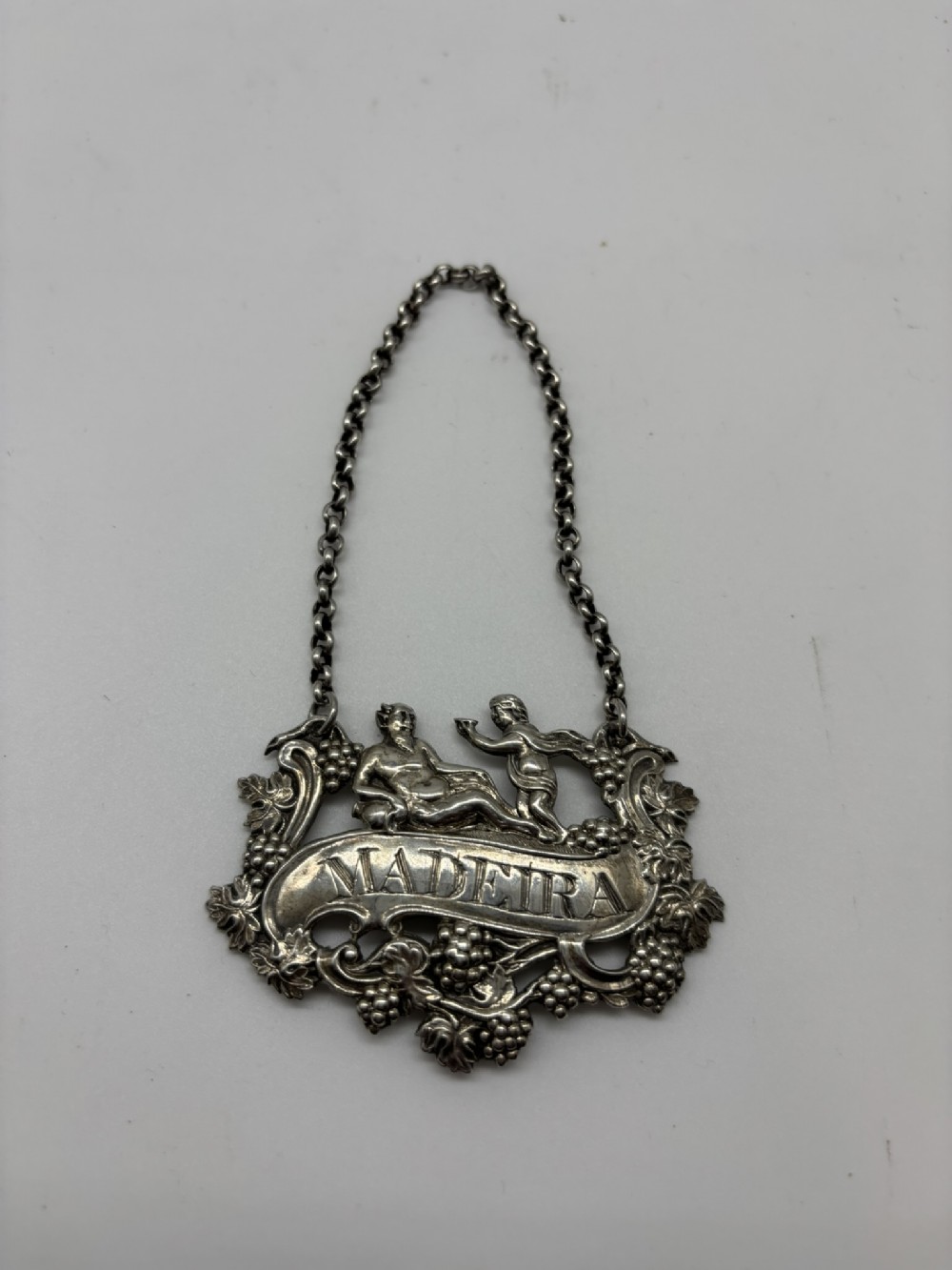 A Georgian sterling silver Madeira decanter label featuring relief carvings of cherubs and grapevines suspended from a silver chain.