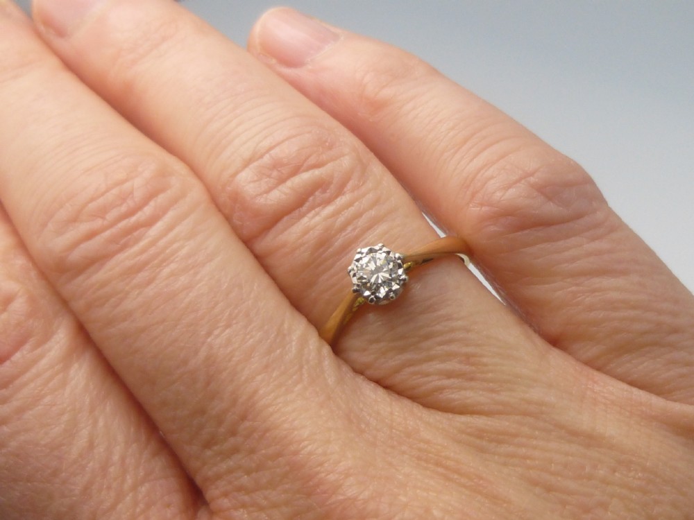 A yellow gold solitaire ring featuring a single round brilliant-cut diamond held in a white metal claw setting. View VI