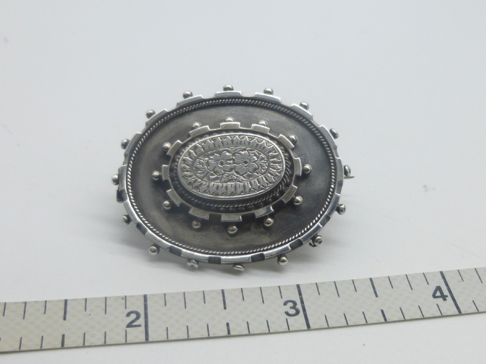 An oval Victorian silver locket brooch featuring an engraved floral centre, rope-twist edging, and a beaded border. View IV