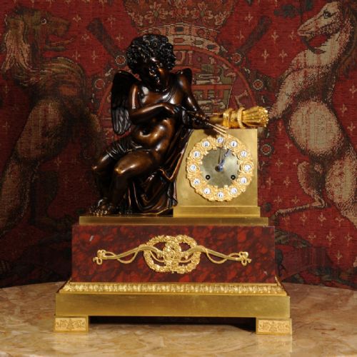 fig 1. A French Empire style dark bronze and gilt ormolu mantel clock featuring a seated Cupid beside the clock dial, supported on a red marble base.