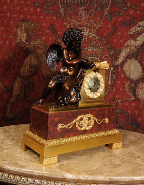 fig 4. A French Empire style dark bronze and gilt ormolu mantel clock featuring a seated Cupid beside the clock dial, supported on a red marble base.