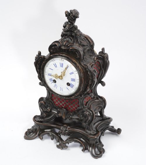 fig 1. An ornate dark bronze Rococo mantel clock featuring a cherub finial, scrolling rocaille design on a matching plinth, a white enamel dial with Roman numerals, and red textile backing panels.