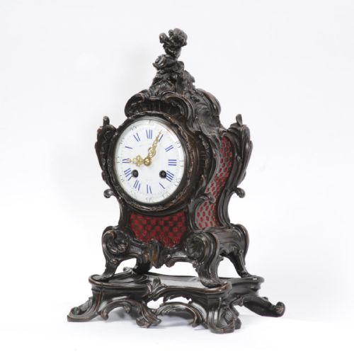 fig 2. An ornate dark bronze Rococo mantel clock featuring a cherub finial, scrolling rocaille design on a matching plinth, a white enamel dial with Roman numerals, and red textile backing panels.