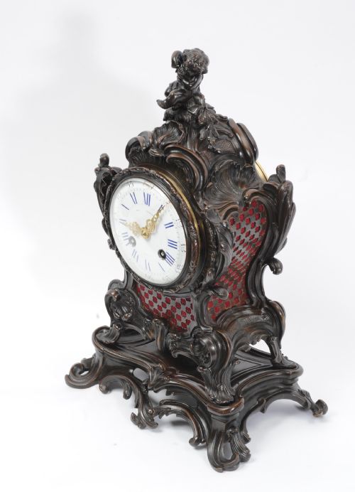fig 3. An ornate dark bronze Rococo mantel clock featuring a cherub finial, scrolling rocaille design on a matching plinth, a white enamel dial with Roman numerals, and red textile backing panels.