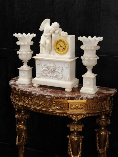 fig 1. A three-piece white alabaster clock set featuring a central timepiece surmounted by a sculpted Cupid figure after Falconet, flanked by two matching carved urns.