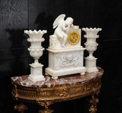fig 2. A three-piece white alabaster clock set featuring a central timepiece surmounted by a sculpted Cupid figure after Falconet, flanked by two matching carved urns.