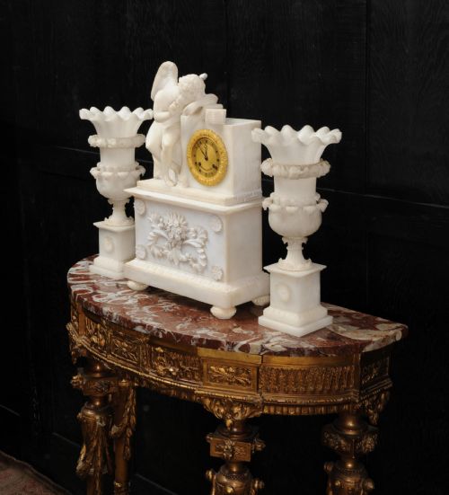 fig 3. A three-piece white alabaster clock set featuring a central timepiece surmounted by a sculpted Cupid figure after Falconet, flanked by two matching carved urns.