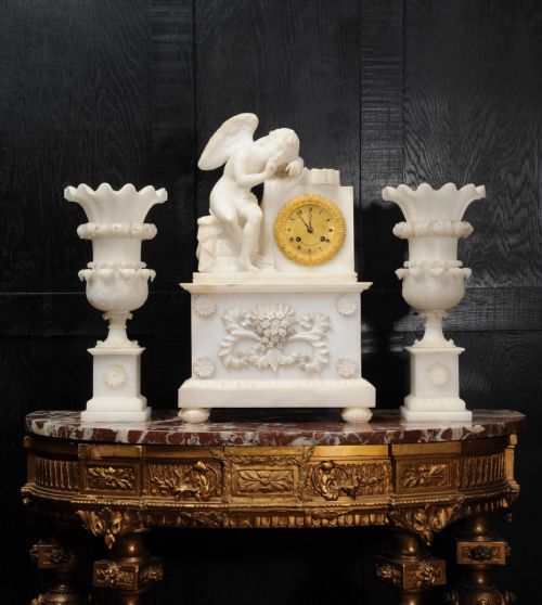 fig 6. A three-piece white alabaster clock set featuring a central timepiece surmounted by a sculpted Cupid figure after Falconet, flanked by two matching carved urns.