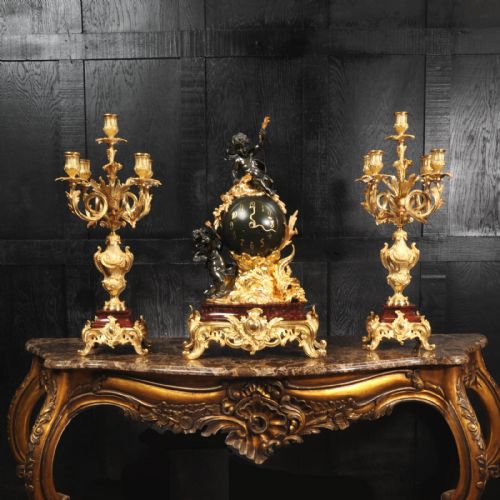 fig 1. An antique French Louis XV style clock garniture set featuring a central gilt bronze and dark bronze cherub clock on a red marble base, flanked by matching multi-arm gilt candelabra.