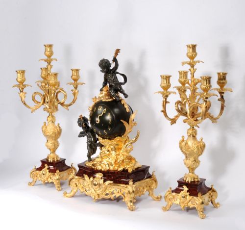 fig 2. An antique French Louis XV style clock garniture set featuring a central gilt bronze and dark bronze cherub clock on a red marble base, flanked by matching multi-arm gilt candelabra.