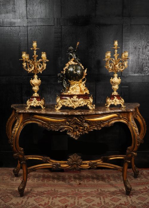 fig 3. An antique French Louis XV style clock garniture set featuring a central gilt bronze and dark bronze cherub clock on a red marble base, flanked by matching multi-arm gilt candelabra.