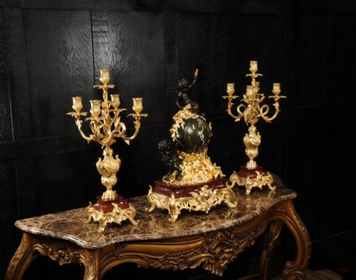 fig 4. An antique French Louis XV style clock garniture set featuring a central gilt bronze and dark bronze cherub clock on a red marble base, flanked by matching multi-arm gilt candelabra.