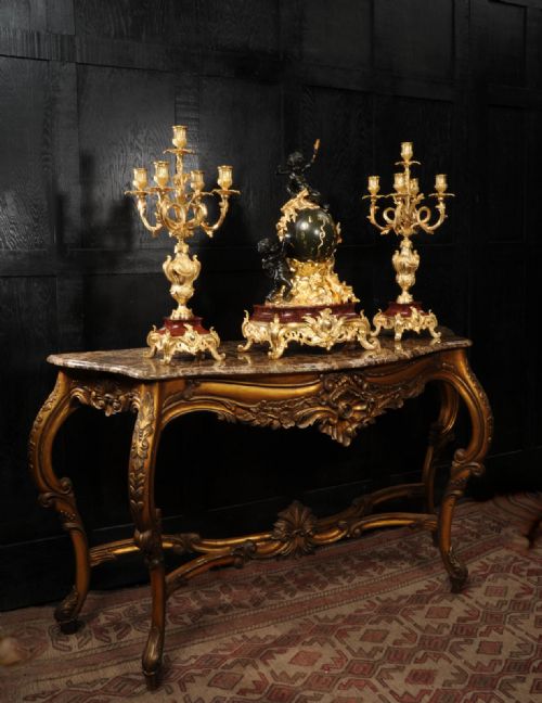 fig 5. An antique French Louis XV style clock garniture set featuring a central gilt bronze and dark bronze cherub clock on a red marble base, flanked by matching multi-arm gilt candelabra.