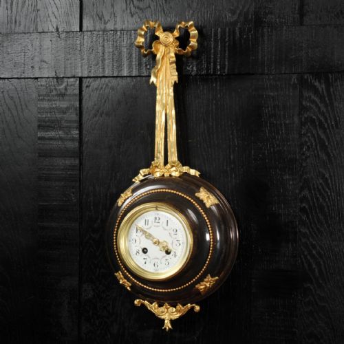 fig 1. A circular wall clock with a white enamel dial, framed in dark bronze with gold ormolu foliate mounts and topped with a gilded ribbon bow.
