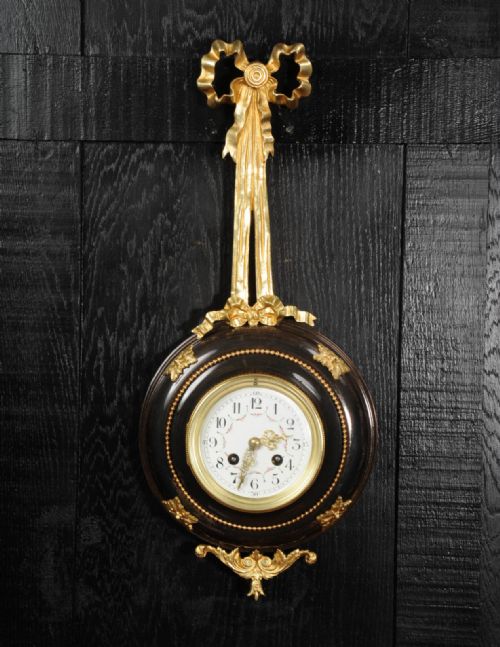 fig 2. A circular wall clock with a white enamel dial, framed in dark bronze with gold ormolu foliate mounts and topped with a gilded ribbon bow.
