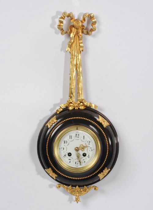 fig 3. A circular wall clock with a white enamel dial, framed in dark bronze with gold ormolu foliate mounts and topped with a gilded ribbon bow.