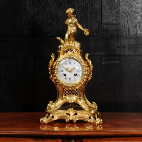 fig 1. A French Rococo gilt bronze mantel clock featuring a cupid figure at the top, scrolling foliage, and a white enamel dial with Roman numerals.