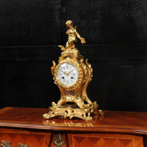 fig 2. A French Rococo gilt bronze mantel clock featuring a cupid figure at the top, scrolling foliage, and a white enamel dial with Roman numerals.