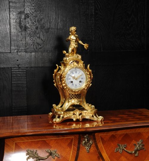 fig 3. A French Rococo gilt bronze mantel clock featuring a cupid figure at the top, scrolling foliage, and a white enamel dial with Roman numerals.